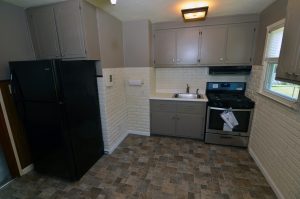 Older kitchen in Groveville Hamilton Township NJ with gray cabinetry, painted brick backsplash, black appliances and patterned flooring.