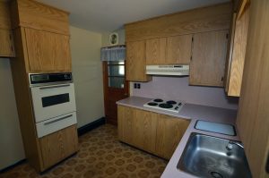 Older kitchen in a New Egypt NJ ranch home with wood cabinetry, pink laminate countertops, patterned flooring and original-style appliances.